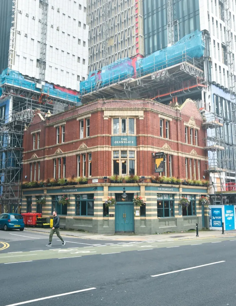 The Deansgate, a red brick Victorian pub with green tiles and hanging baskets, on a corner of Deansgate in Manchester with new towers under scaffolding behind it.