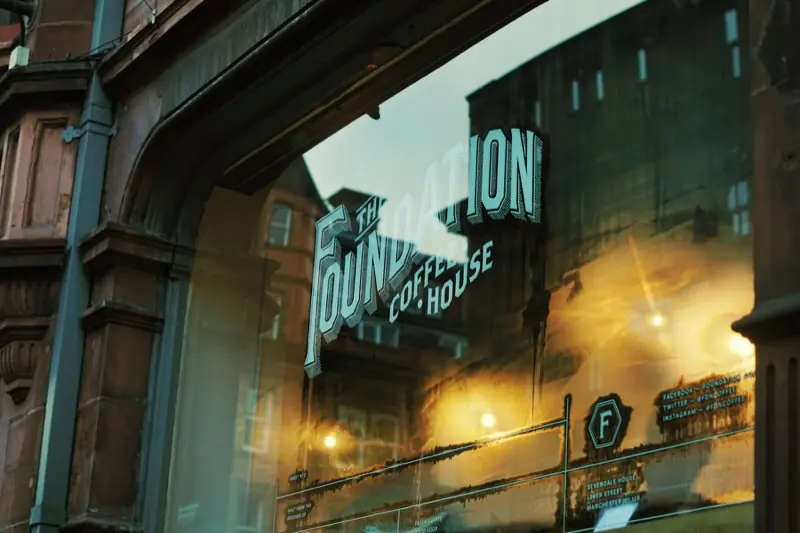 The Foundation Coffee House painted on the window of Sevendale House on Lever Street, in Manchester's Northern Quarter, with warm lamps inside and red brick buildings reflected in the glass.