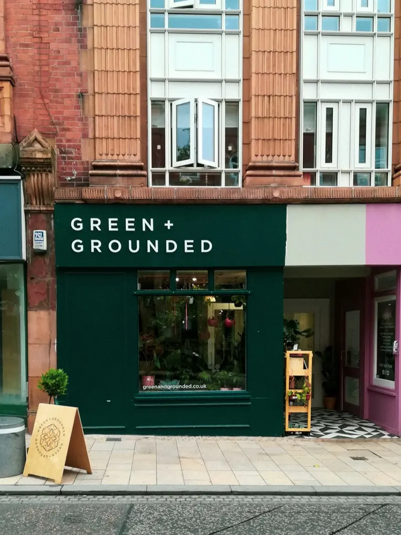Green + Grounded, a plant shop in Altrincham with a dark green front and a window full of plants, a wooden A board and a clipped bay tree in a pot on the pavement.