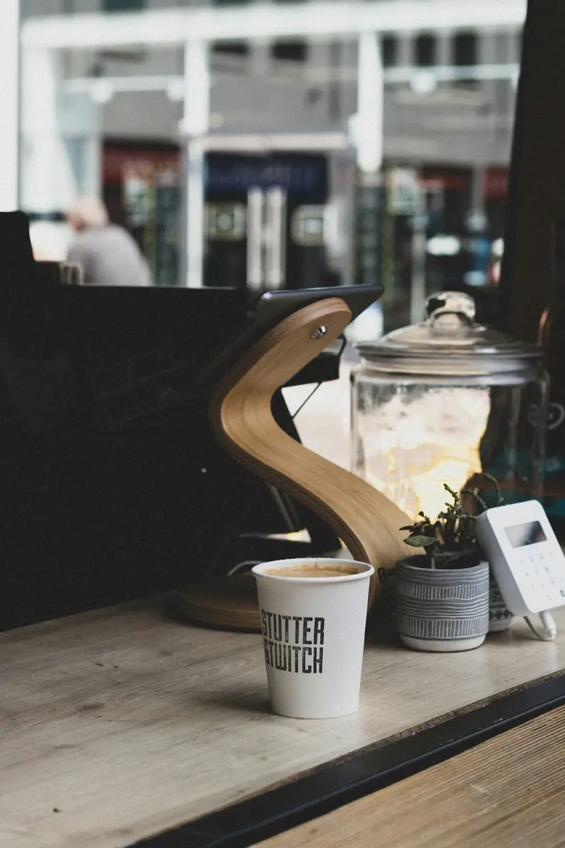 A Stutter & Twitch paper cup of coffee on a wooden counter at the coffee and record bar in Altrincham, next to a small plant, a glass jar and a card reader.